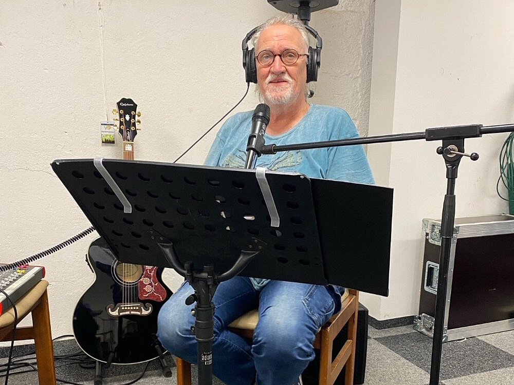 Ein Bandmitglied sitzt vor einem Ständer mit Text, hat ein Mikrofon vor sich und schaut in die Kamera.ch und 