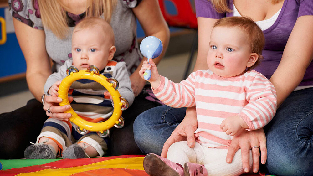 Zwei Babys sitzen auf dem Boden, jedes mit einem Erwachsenem im Hintergrund. Die Babys halten Spielzeuge in den Händen. 