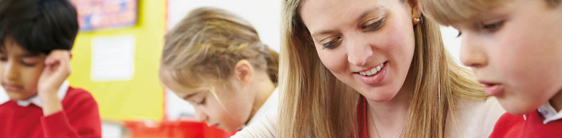 Pädagogische Fachkraft unterstützt einen Schüler im Klassenzimmer beim Lernen, im Hintergrund sind zwei weitere Schüler zu erkennen.