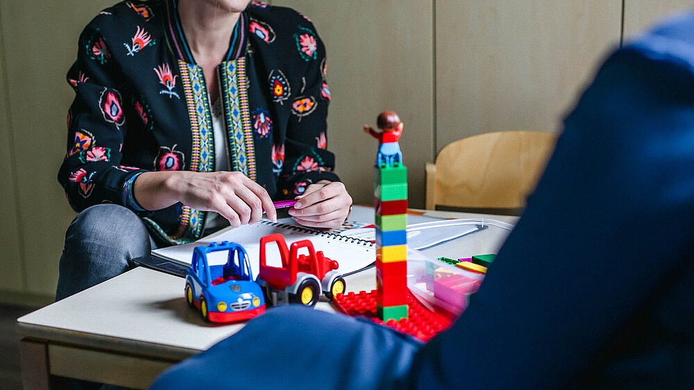 Heilpädagogische Fachkraft sitzt an einem Tisch mit Spielzeugautos und Bauklötzen und ist im Gespräch mit einer weiteren Person. 