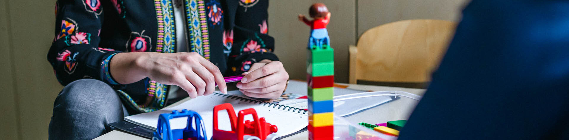 Heilpädagogische Fachkraft sitzt an einem Tisch mit Spielzeugautos und Bauklötzen und ist im Gespräch mit einer weiteren Person. 