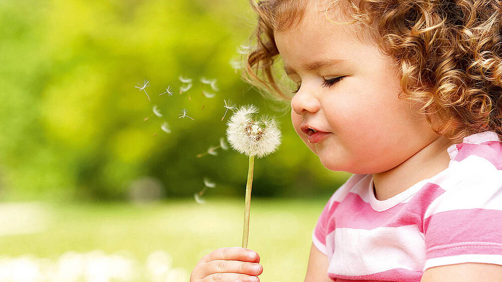 Ein Mädchen sitzt draußen mit einer Pusteblume in der Hand und pustet die einzelnen Samen weg.