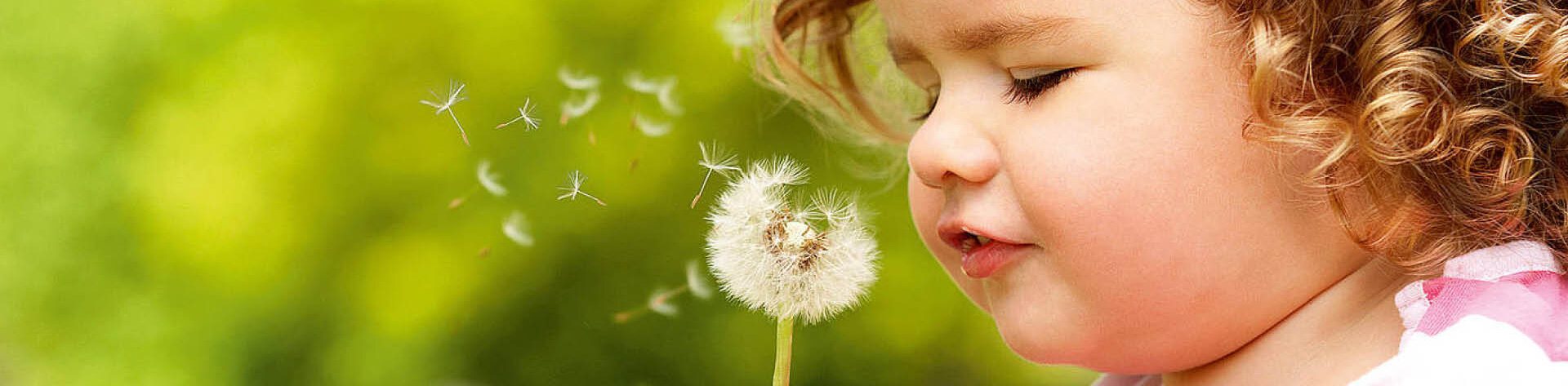 Ein Mädchen sitzt draußen mit einer Pusteblume in der Hand und pustet die einzelnen Samen weg.